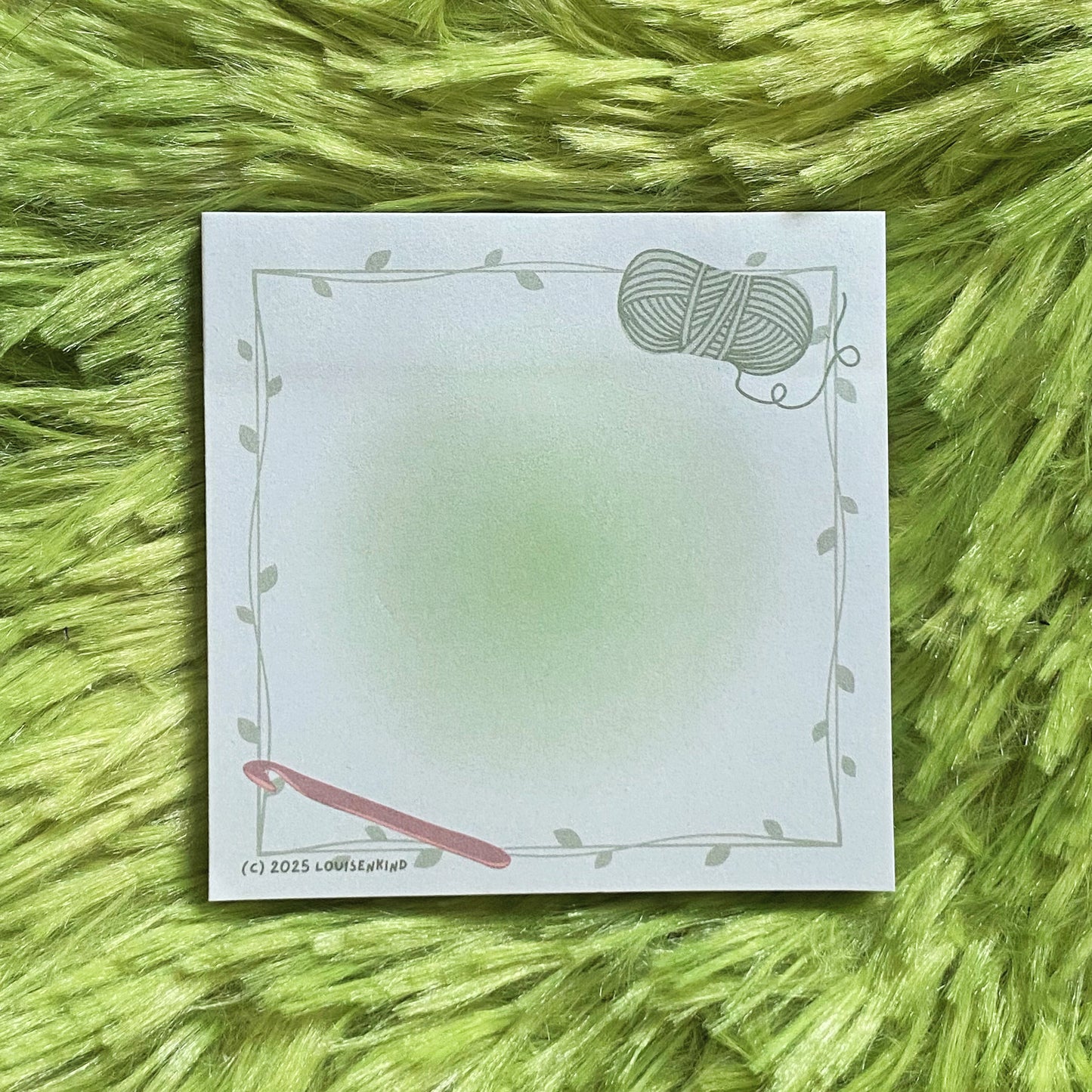 A sticky notepad with crochet hook and a skein of yarn print on it laying on a green fluffy rug