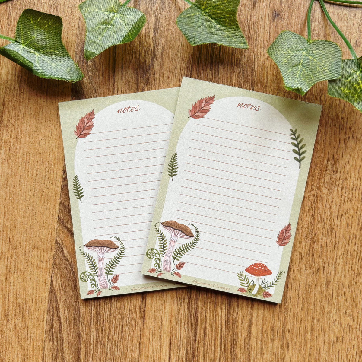 Two mushroom illustrated A7 notepads with 25 lined sheets amanita and chestnut mushrooms with ferns and autumn leaves notes sage green border on wooden table