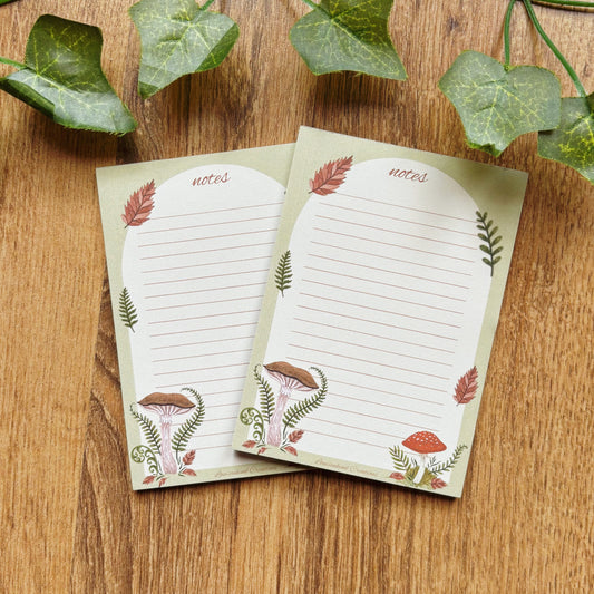 Two mushroom illustrated A7 notepads with 25 lined sheets amanita and chestnut mushrooms with ferns and autumn leaves notes sage green border on wooden table