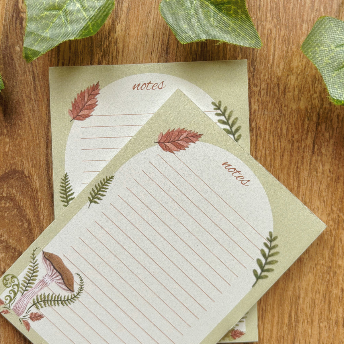Two decorative lined note cards with a woodland theme, featuring illustrations of a mushroom, ferns, and autumn leaves, displayed on a wooden surface with green ivy leaves.